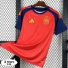 Red Spain national team jersey with yellow pinstripes and navy-blue sleeves, Adidas logo, crest with two stars, hanging on a wooden hanger against a metal grid background.