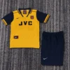 Arsenal yellow away soccer jersey with navy sleeves and JVC sponsor, paired with navy shorts on a gray carpet.