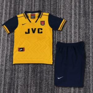 Arsenal yellow away soccer jersey with navy sleeves and JVC sponsor, paired with navy shorts on a gray carpet.
