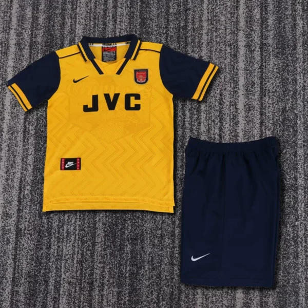 Arsenal yellow away soccer jersey with navy sleeves and JVC sponsor, paired with navy shorts on a gray carpet.