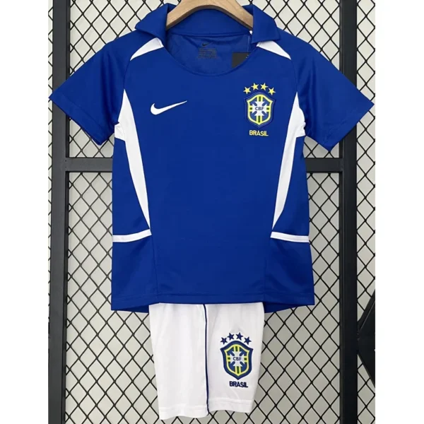 Blue Brazil national team jersey with white accents and crest, paired with white shorts featuring the Brazilian badge, hanging on a hanger in front of a fence.