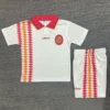 White Spain soccer jersey with red collar, crest, and red–orange diamond stripe on the left; matching white shorts with the same stripe down the side.