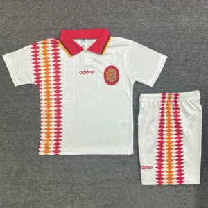 White Spain soccer jersey with red collar, crest, and red–orange diamond stripe on the left; matching white shorts with the same stripe down the side.