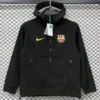 Black zip-up jacket with FC Barcelona crest and yellow Nike swoosh on chest, hanging on a display hanger with tags attached.