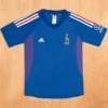 Blue Adidas France national team soccer jersey laid flat; features crest with rooster and star, white Adidas logo, and red-white-blue shoulder stripes on a wooden floor background.