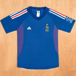 Blue Adidas France national team soccer jersey laid flat; features crest with rooster and star, white Adidas logo, and red-white-blue shoulder stripes on a wooden floor background.