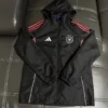 Germany 2026/27 Black Windbreaker - Front View - KFCool – Football Jerseys & Soccer Kits Store Black hooded windbreaker with red stripes on the shoulders, white Adidas logo, and a Germany crest, laid on a dark leather couch.