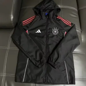 Germany 2026/27 Black Windbreaker - Front View Black hooded windbreaker with red stripes on the shoulders, white Adidas logo, and a Germany crest, laid on a dark leather couch.