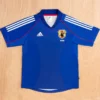 Blue Japan national football jersey laid flat, featuring Adidas logo and JFA crest with three white stripes on the sleeves.