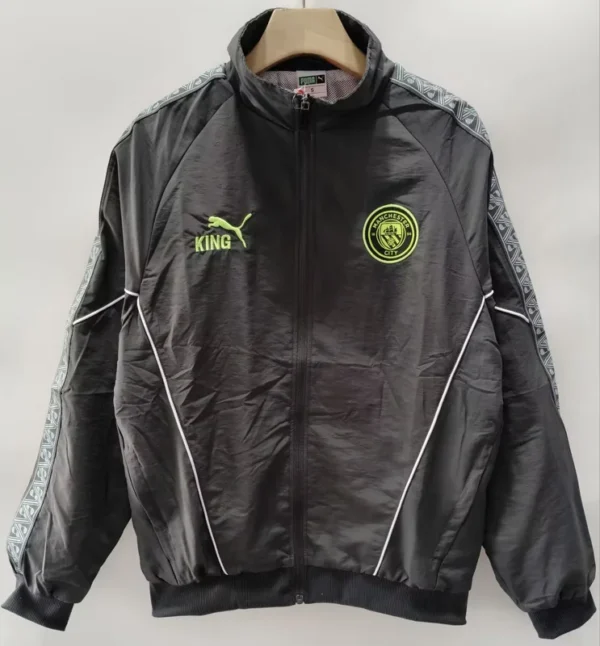 Manchester City 2026/27 Windbreaker - Front View - KFCool – Football Jerseys & Soccer Kits Store Black Manchester City track jacket on a hanger, with neon crest on the left chest, neon Puma King logo on the right, white piping, and a patterned sleeve design.