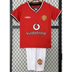 Manchester United 2000/02 Retro Home Kids Kit Fan Version - Front View Red Manchester United football jersey with Vodafone sponsor, white-trim V-neck, crest on the left chest, paired with white shorts showing the club badge, hanging against a fence.