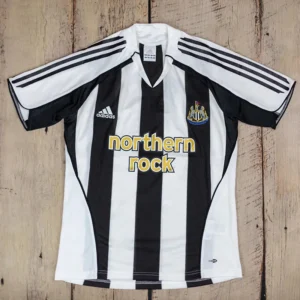 Newcastle United 2005/06 Retro Home Jersey Fan Version - Front View Black and white striped Adidas Newcastle United home jersey with Northern Rock sponsor laid flat on a wooden surface.