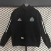Black Adidas hoodie featuring a Newcastle United crest on the chest, white sleeve stripes, front pocket, hanging on a display against a grid backdrop.