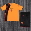 Roma 2005/06 Retro Home Kids Kit Fan Version - Front View - KFCool – Football Jerseys & Soccer Kits Store Orange Diadora soccer jersey with black diagonal stripe and team crest, paired with black shorts featuring a crest on a gray carpeted background.