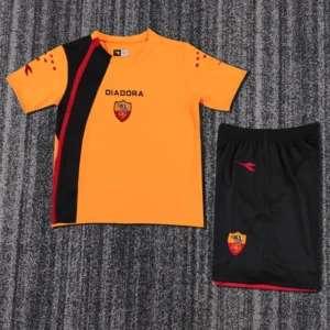 Roma 2005/06 Retro Home Kids Kit Fan Version - Front View Orange Diadora soccer jersey with black diagonal stripe and team crest, paired with black shorts featuring a crest on a gray carpeted background.
