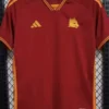 Roma 2023/24 Home Jersey Fan Version - Front View Maroon Adidas sports jersey hanging on a hanger with yellow trim and a wolf crest on the left chest.