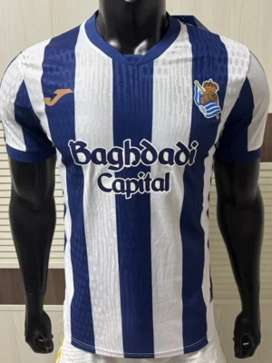 Real Sociedad 2026/27 Home Jersey Player Version - Front View Mannequin wearing a blue and white vertical stripe soccer jersey with the sponsor 'Baghdad Capital' across the chest and a team crest on the left.